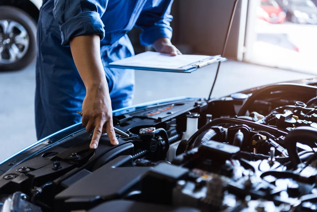 Auto mechanic in blue uniform holding a clipboard and pointing to a part of the engine while performing an inspection on a car.