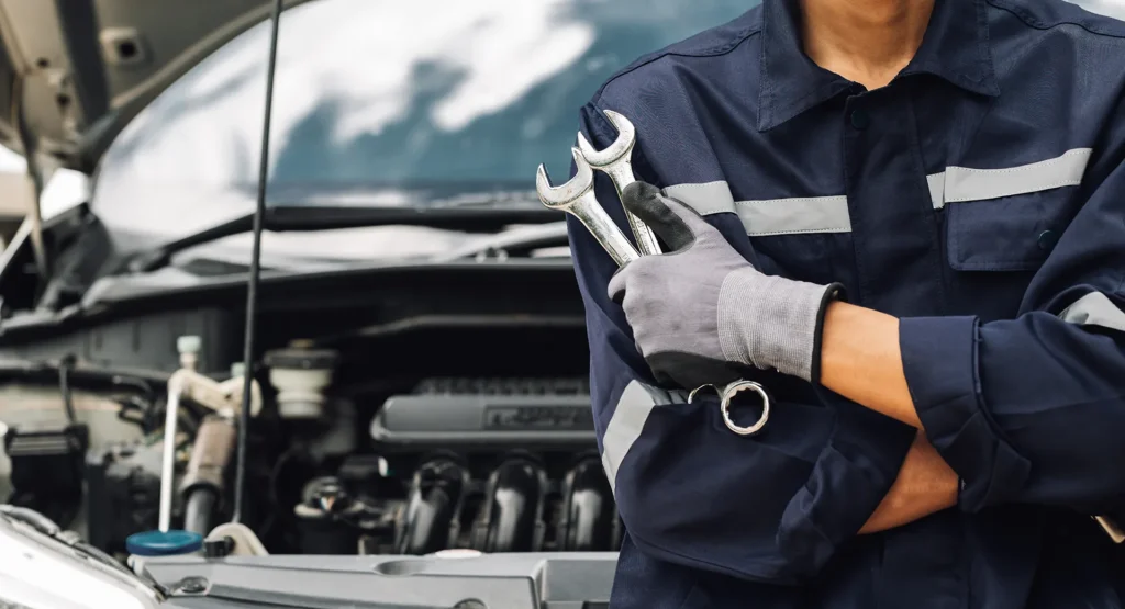 Close-up of a car mechanic in a dark blue uniform and gloves holding two silver combination wrenches next to an open car hood.