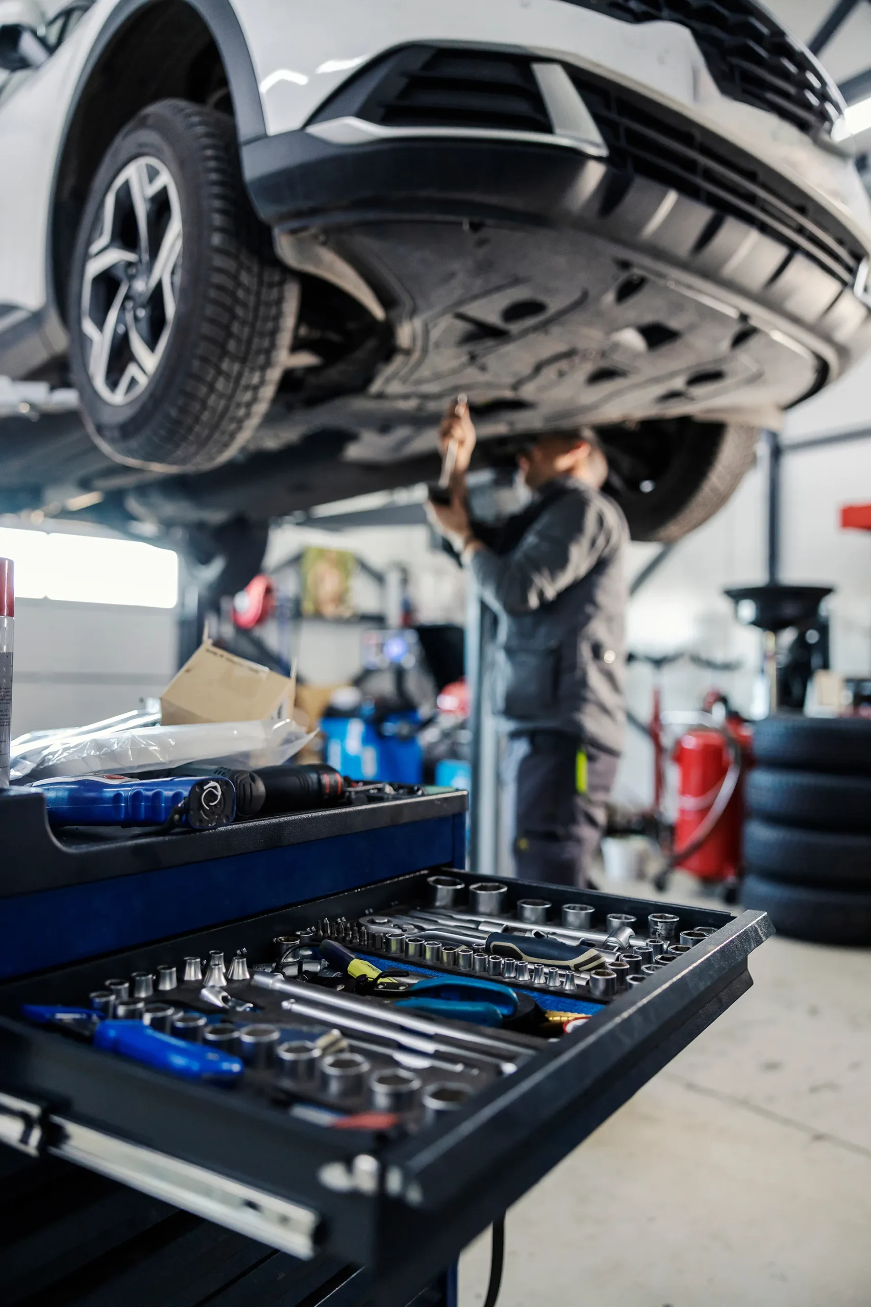 Open toolbox with socket wrenches and automotive tools in the foreground, with a blurry mechanic working under a car lifted on a hydraulic hoist in the background.